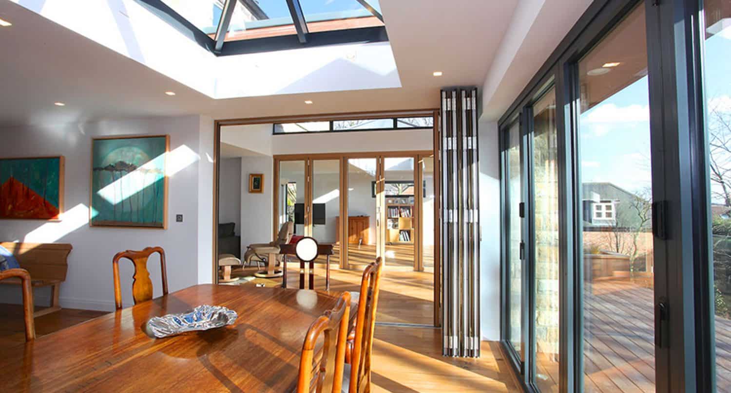 Bright modern dining area with wooden table and chairs, large glass doors open to a deck, and skylights letting in sunlight. Contemporary art hangs on white walls, creating an open, airy feel.