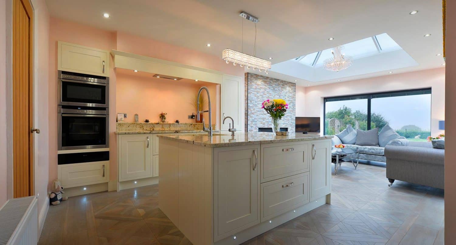 Modern kitchen with cream cabinets, an island sink, built-in oven, and a vase of flowers. Open-plan layout connects to a cozy living area with sofas and large windows letting in natural light.