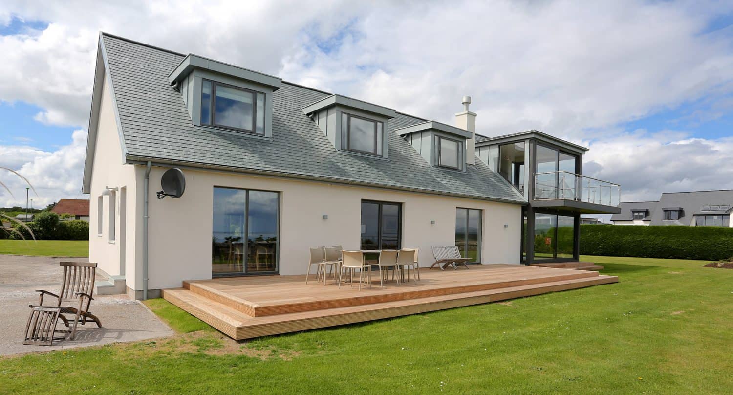 Modern two-story house with gray roof, large windows, and a spacious wooden deck with outdoor furniture, surrounded by a green lawn and clear sky with some clouds.