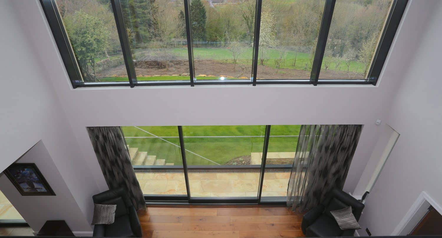 A view from an upper floor looking through large floor-to-ceiling windows onto a green garden, with two black armchairs and patterned curtains below.