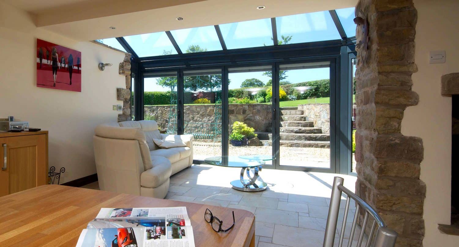 A bright sunroom with glass ceiling and sliding doors opens to a stone terrace and garden. Inside are a cream sofa, a small glass table, and a wooden table with a magazine and eyeglasses on it.