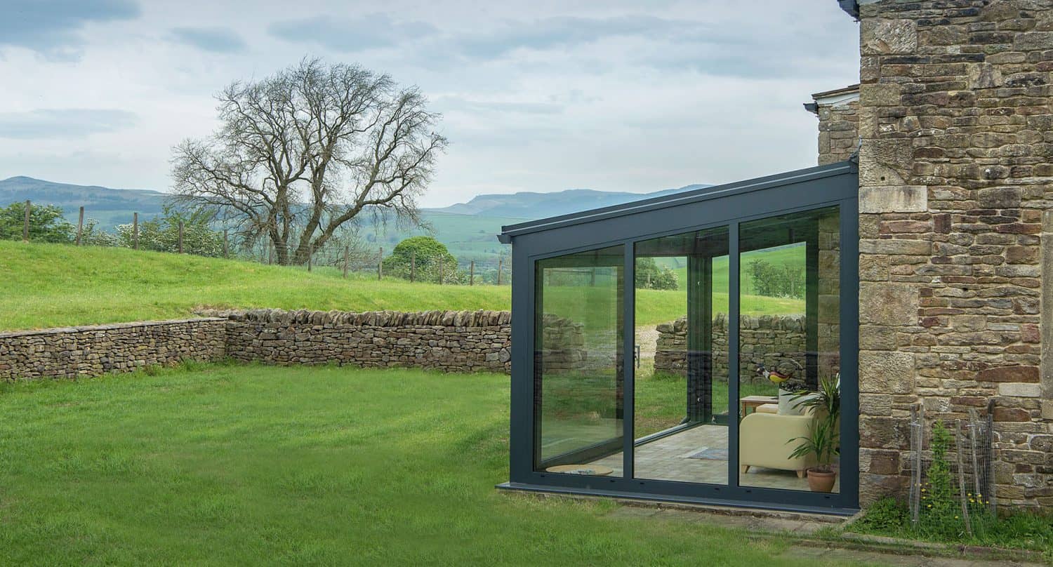 A modern glass-walled sunroom with a sofa and potted plant is attached to a stone house, overlooking a grassy yard, stone wall, and a scenic landscape with hills and a leafless tree.