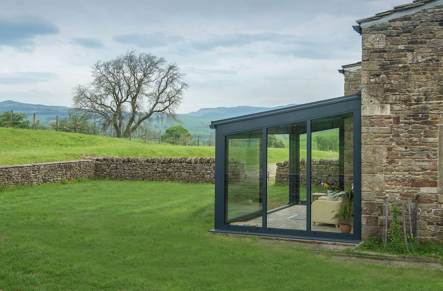 A modern glass-walled sunroom with a sofa and potted plant is attached to a stone house, overlooking a grassy yard, stone wall, and a scenic landscape with hills and a leafless tree.