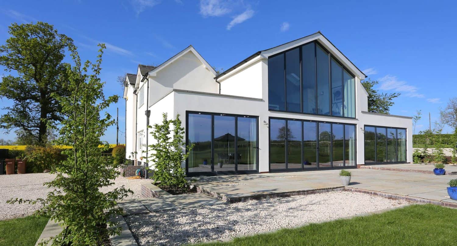 Modern white house with large floor-to-ceiling windows, surrounded by a landscaped garden with young trees, gravel paths, and a stone patio, set against a bright blue sky.