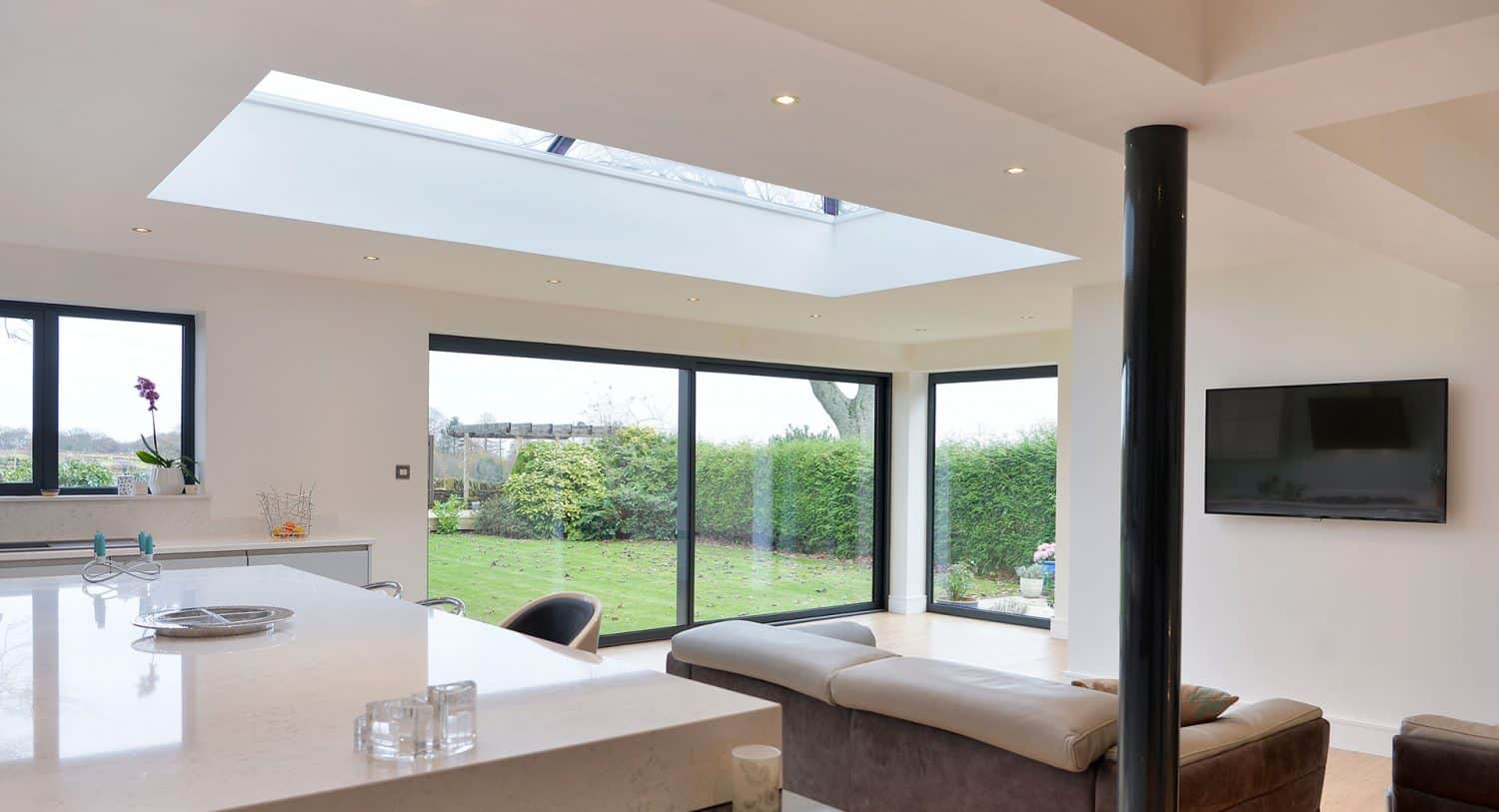 Modern, bright living space with large sliding glass doors, a skylight, white walls, a corner sofa, wall-mounted TV, and sleek bifold doors opening to views of a green garden outside.