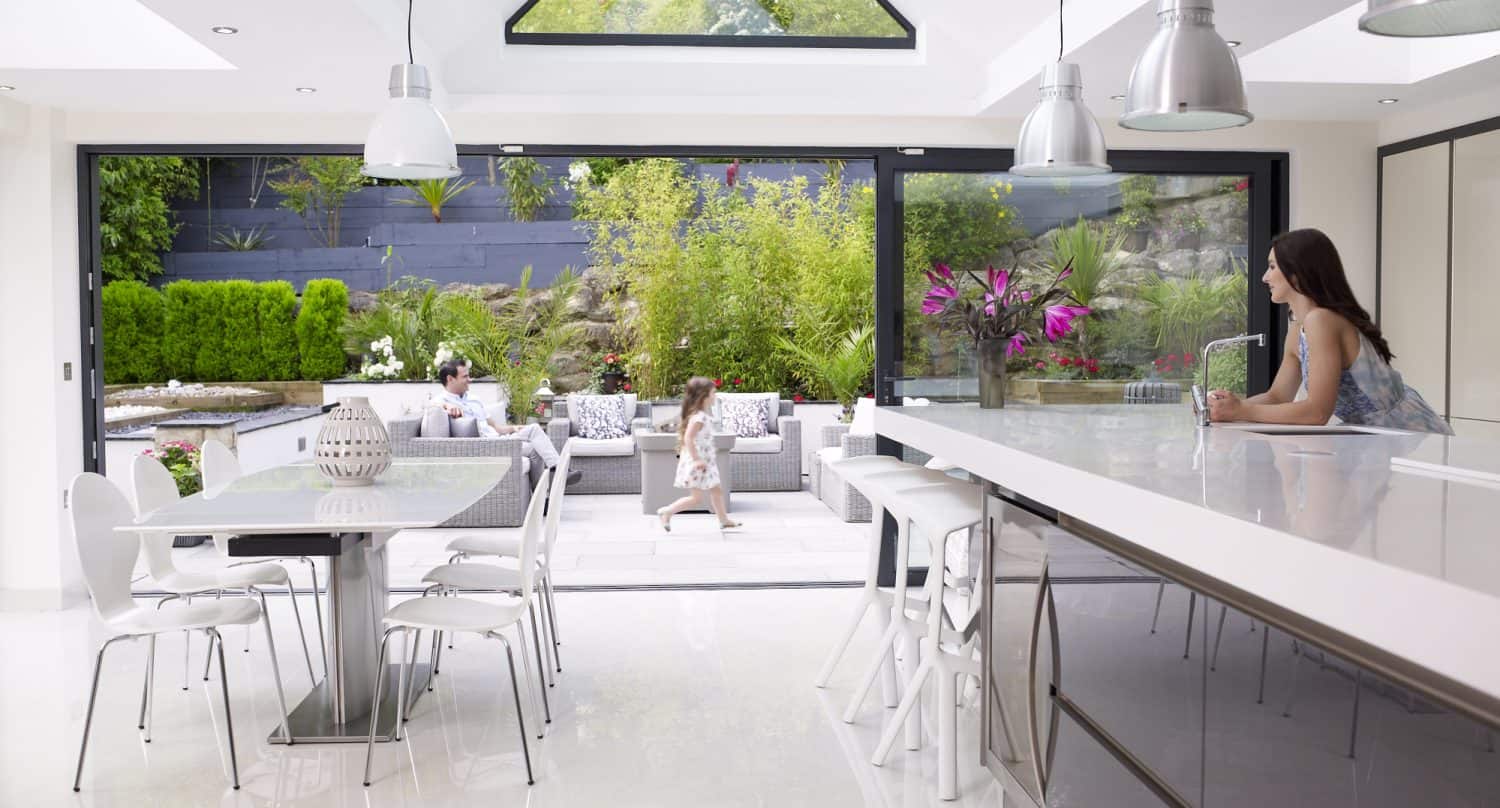 Modern, bright kitchen with white furniture and large bifold doors opening to a lush garden patio. A woman leans on the kitchen island, while two people relax on an outdoor sofa and a child walks nearby.