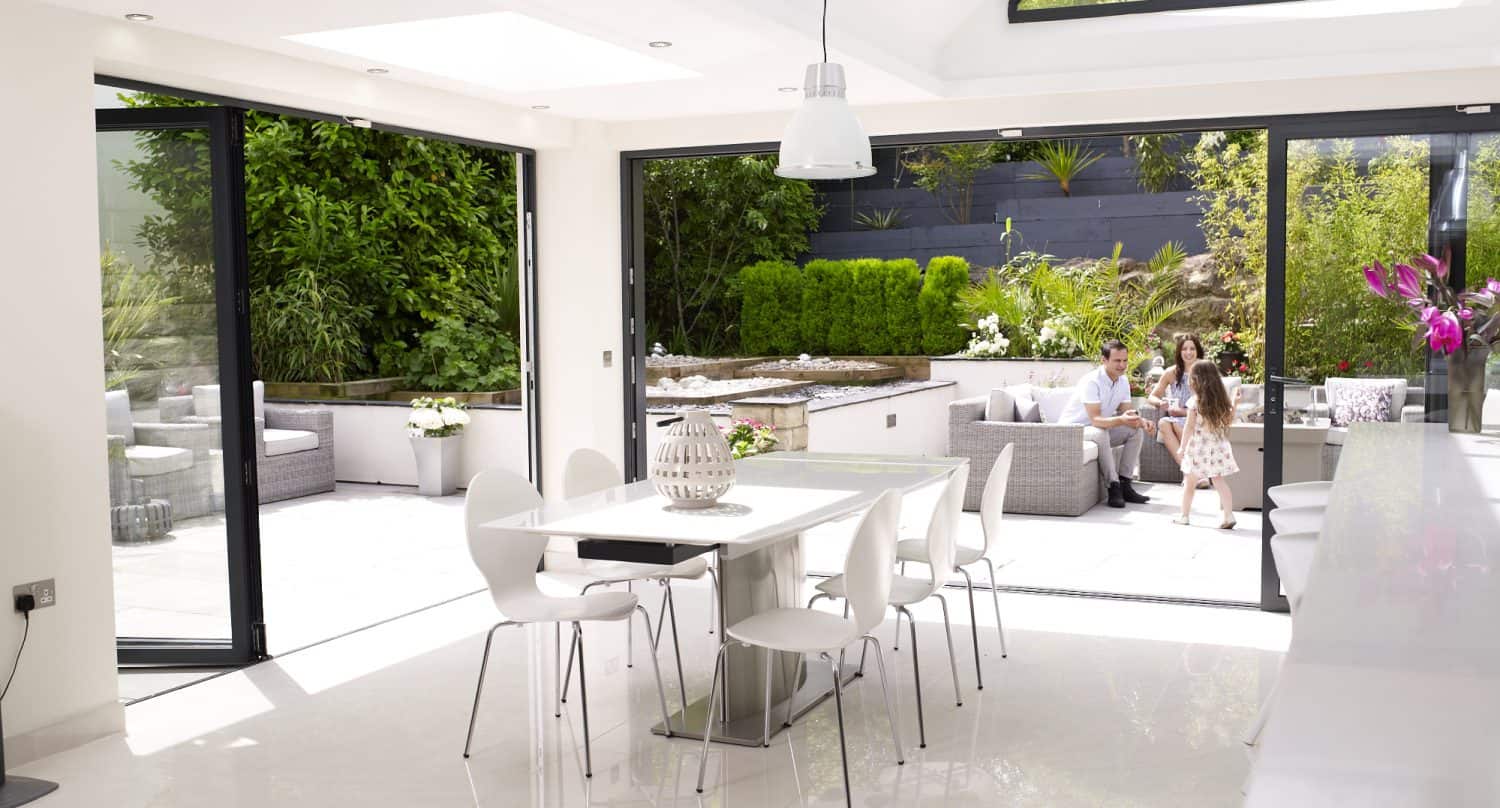 Modern, bright dining area with white furniture and open bifold doors leading to a lush patio garden where two adults and a child are sitting and walking. Sunlight fills the space, highlighting the seamless indoor-outdoor living.