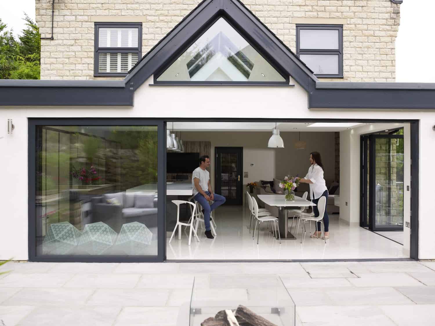 A man and a woman are talking in a modern, open-plan kitchen and dining area with large sliding doors. The space is bright with white walls, contemporary furniture, and views of a stone patio outside.