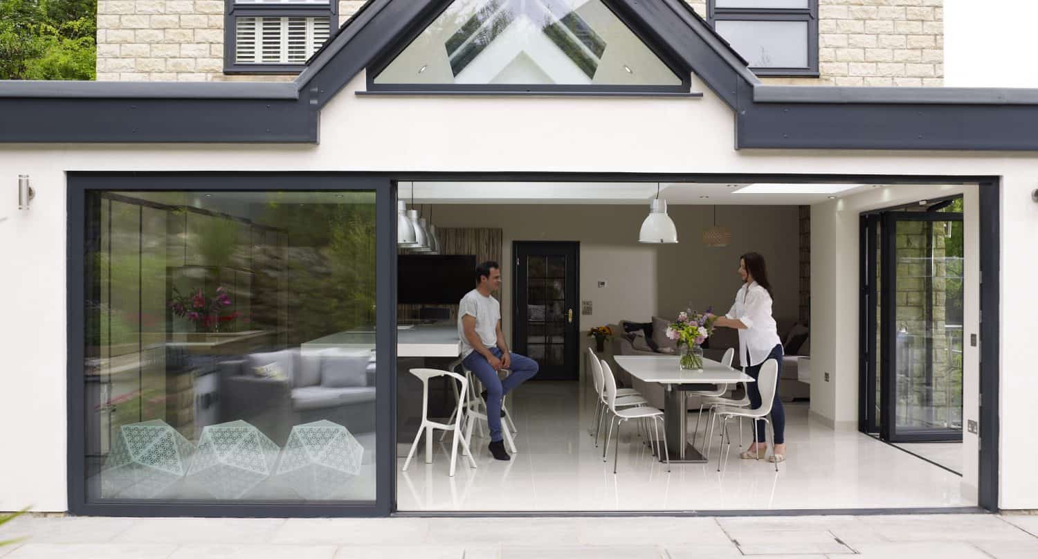 A man and a woman are talking in a modern, open-plan kitchen and dining area with large sliding doors. The space is bright with white walls, contemporary furniture, and views of a stone patio outside.