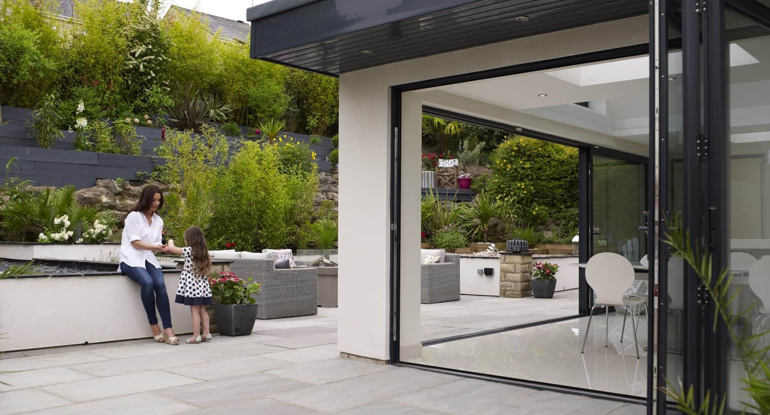 A woman and a young girl sit together on a patio bench in a modern garden, surrounded by greenery and flowers, with bifold doors open to a bright indoor dining area.