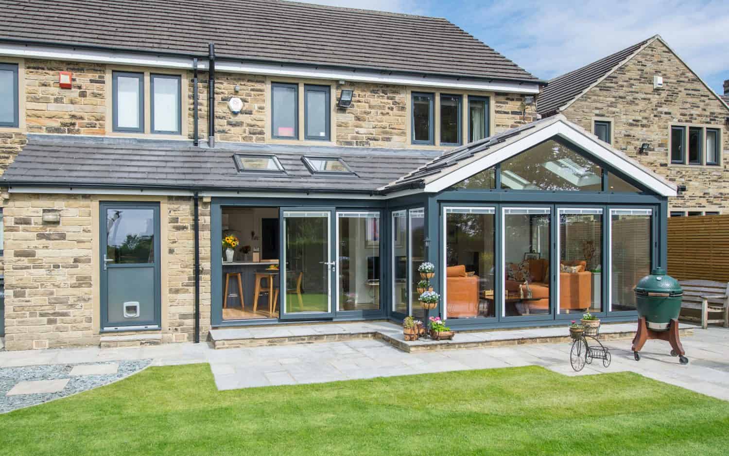 Modern stone house with a glass sunroom extension, featuring large windows and sliding doors opening onto a neatly trimmed lawn and patio with outdoor furniture and a barbecue grill.