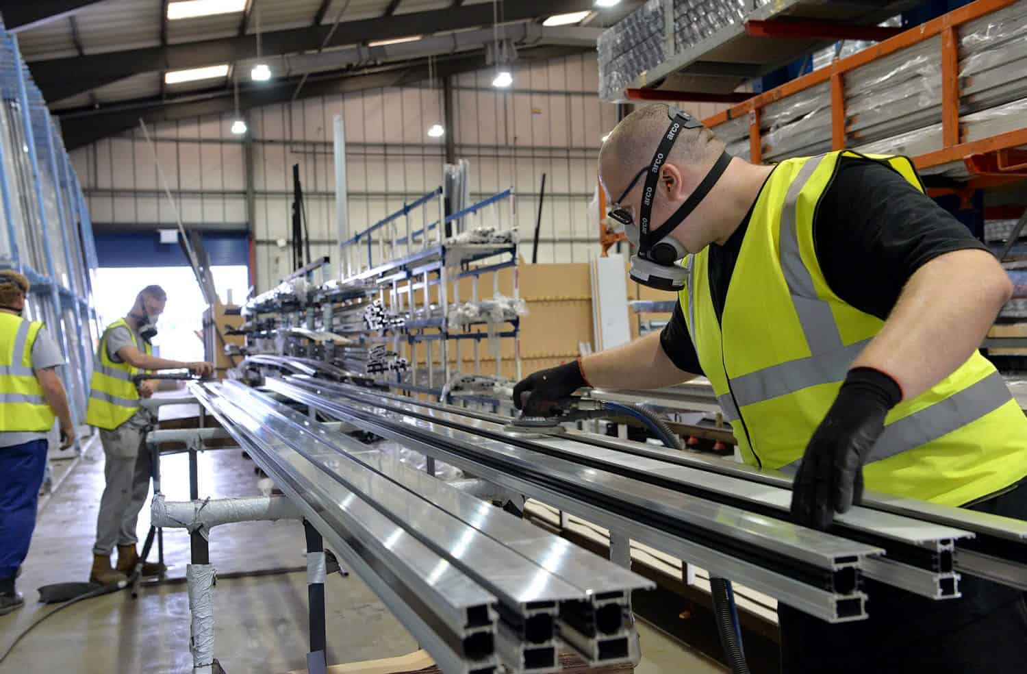 A worker wearing a high-visibility vest, gloves, and a protective mask handles long metal beams in a large, organized factory. Another worker is visible in the background, also working with metal materials.
