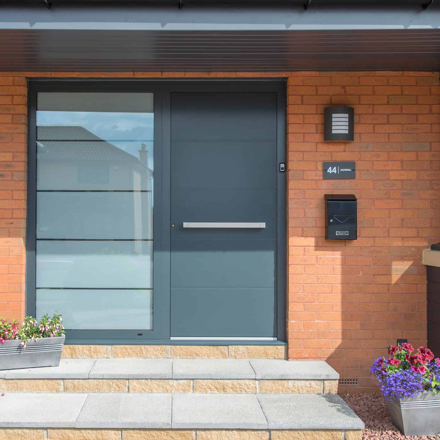 Modern front door with frosted glass panels and a gray mailbox on a brick house. House number 44 is displayed next to the door, with bifold doors nearby and potted flowers on either side of the entrance.
