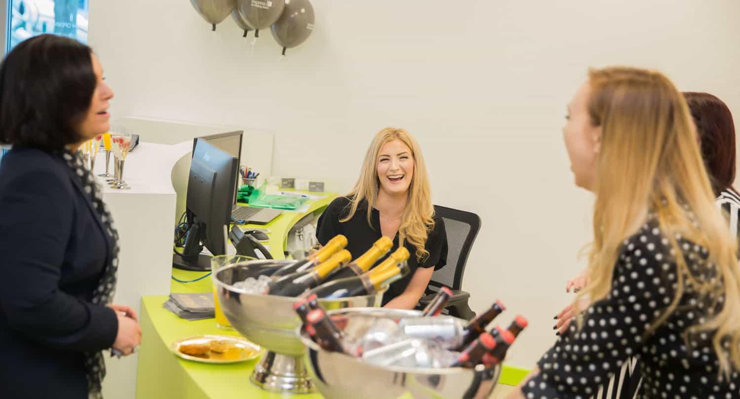 Four women are gathered around a desk with buckets of wine and champagne bottles, smiling and conversing in a bright, modern office with sliding doors. One woman sits at the desk, laughing.