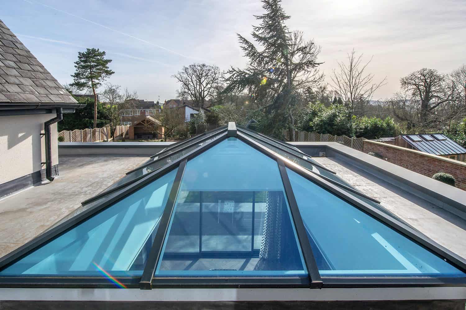 A large glass roof skylight with a triangular design sits atop a flat roof, reflecting the sky and nearby trees, with houses and a tall tree visible in the background.