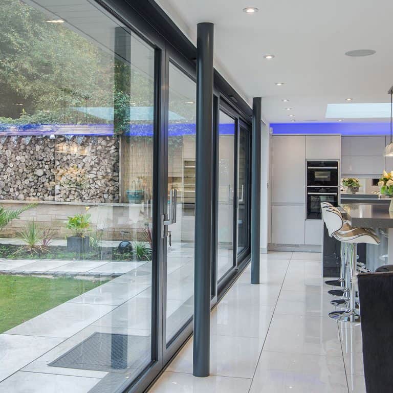 Modern kitchen with glossy white floors and large glass sliding doors opening to a garden. Bar stools line a counter, and stacked firewood is visible outside near greenery and a brick wall.