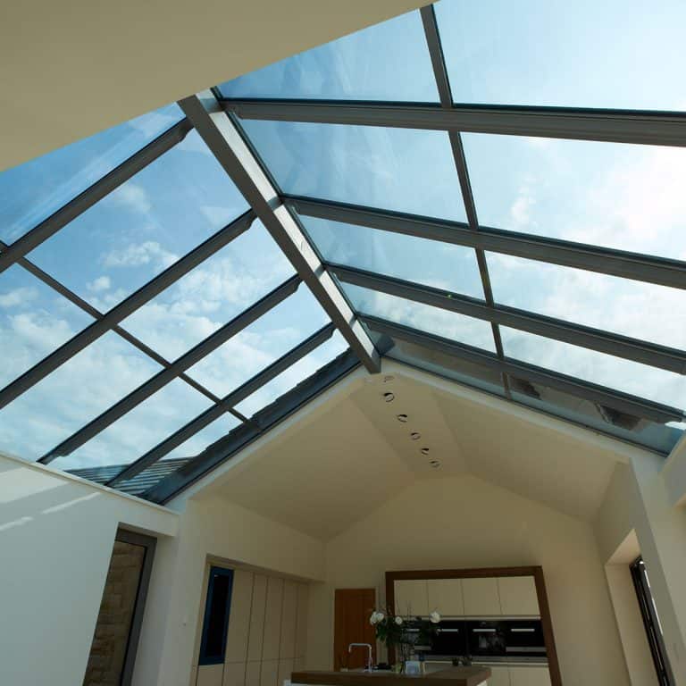 Modern kitchen interior with a high sloped glass ceiling, allowing natural light to flood the room. The clear blue sky and scattered clouds are visible through the glass panels above.