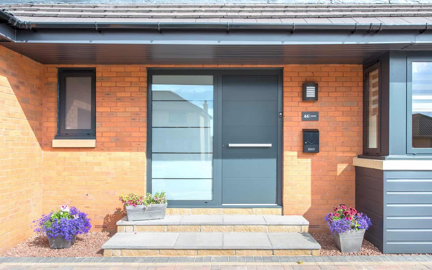Modern aluminium front doors with a large frosted glass panel grace this brick house. Stone steps lead up to the entrance, flanked by potted flowers and small windows, with a black mailbox mounted on the wall.