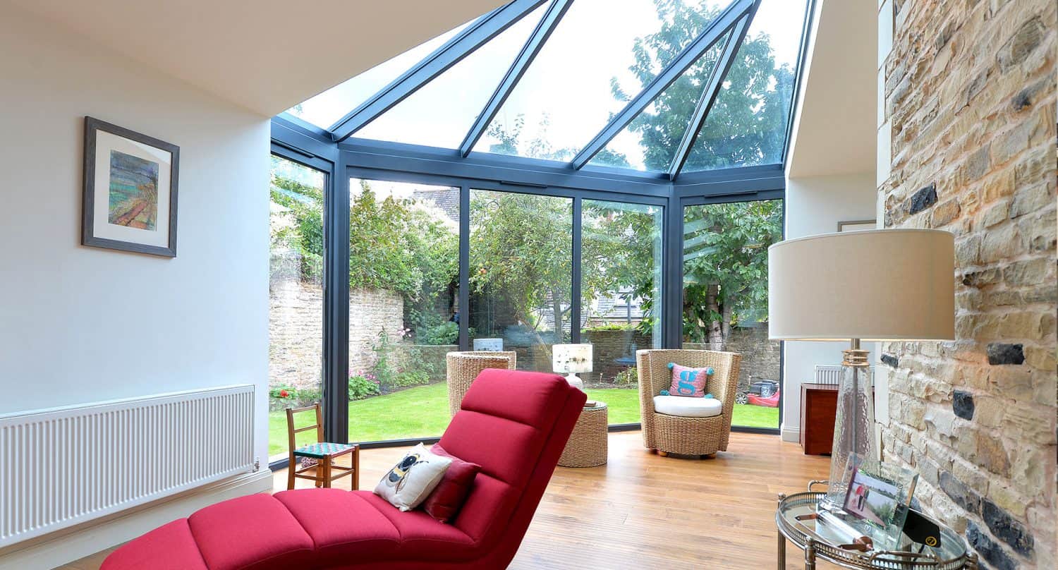 A bright sunroom with a glass ceiling and walls, featuring a red chaise lounge, wicker chairs, a floor lamp, wooden flooring, and a stone accent wall, overlooking a green garden outside.