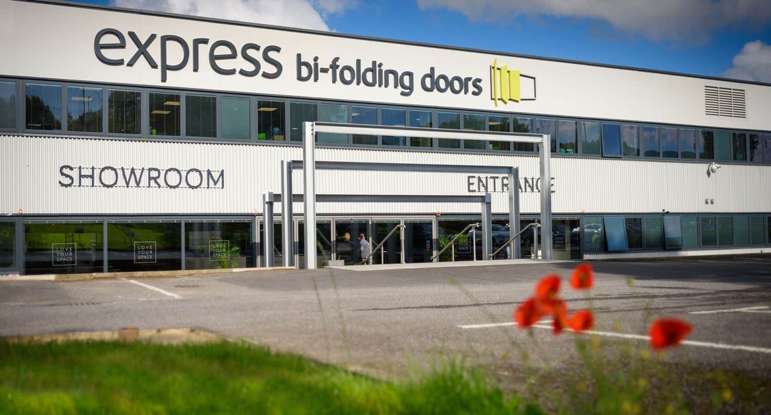 A modern building with large windows and signs reading express bi-folding doors, SHOWROOM, and ENTRANCE. Red flowers and grass are in the foreground under a partly cloudy sky.