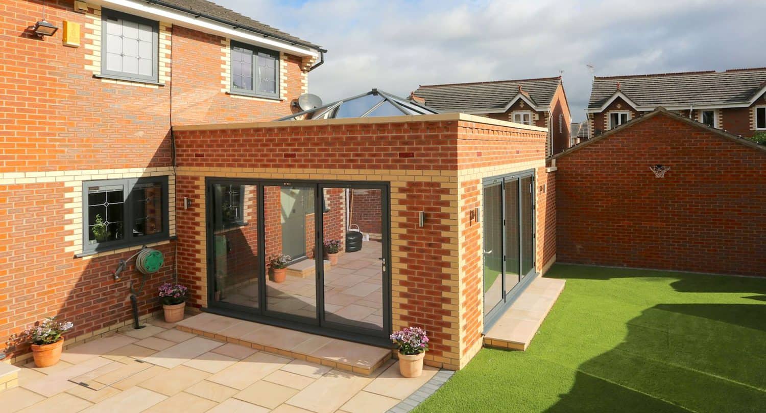Modern brick house with a glass-roofed extension, large sliding glass doors, potted plants, and a patio area leading to artificial grass lawn; neighboring brick houses visible in the background.