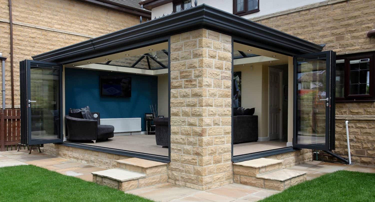 Modern sunroom extension with large glass folding doors, beige stone walls, and a black roof. The space opens to a patio and lawn, with a cozy seating area and blue accent wall inside.