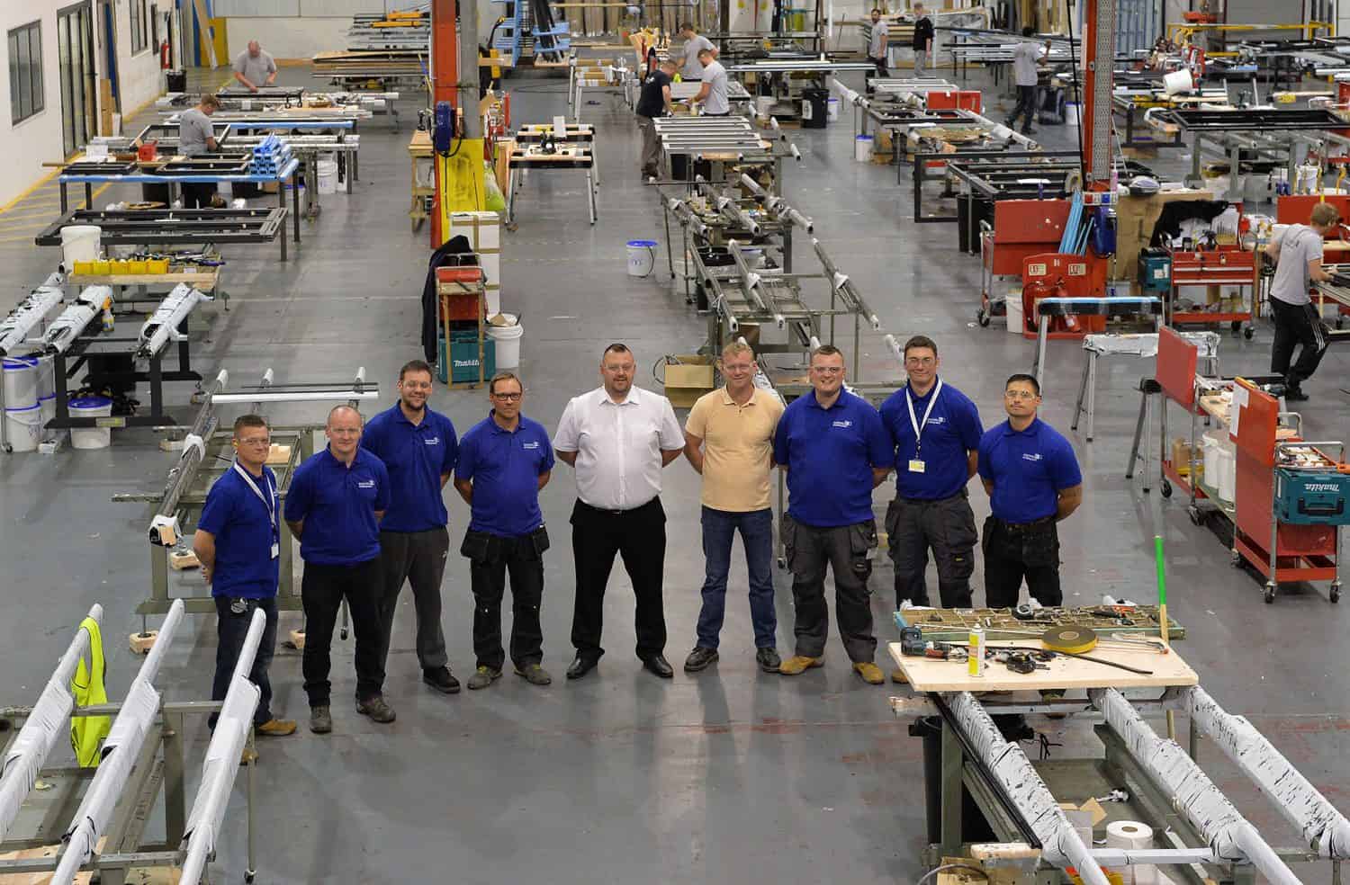 A group of nine men stand together and pose for a photo inside a large, organized factory filled with workstations, tools, and machinery. Other workers are visible in the background working.
