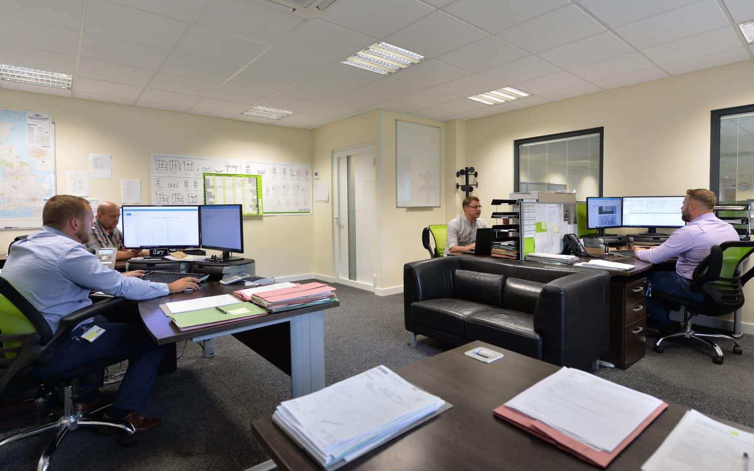 Four men work at desks with computers and paperwork in a modern office. Whiteboards, maps, and documents are on the walls. The office has light-colored walls, carpeted floors, and overhead fluorescent lighting.