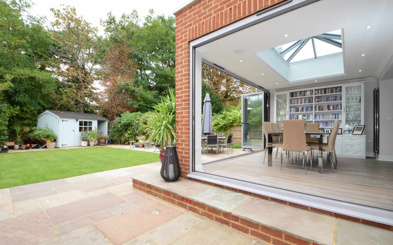 A modern dining area with glass doors opens onto a stone patio and lush garden. There is a lawn, potted plants, outdoor furniture, and a small white shed surrounded by trees in the background.