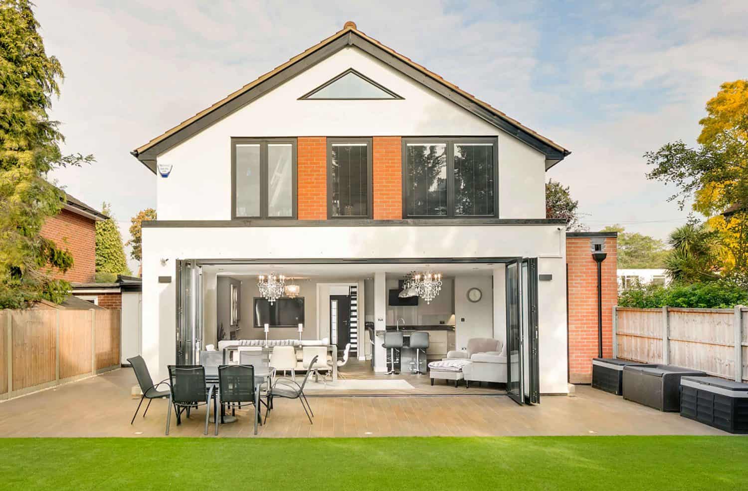 A modern two-story house with large glass doors open to a spacious patio. The patio has outdoor furniture, and the interior features a stylish living and dining area. The yard is covered with artificial grass.