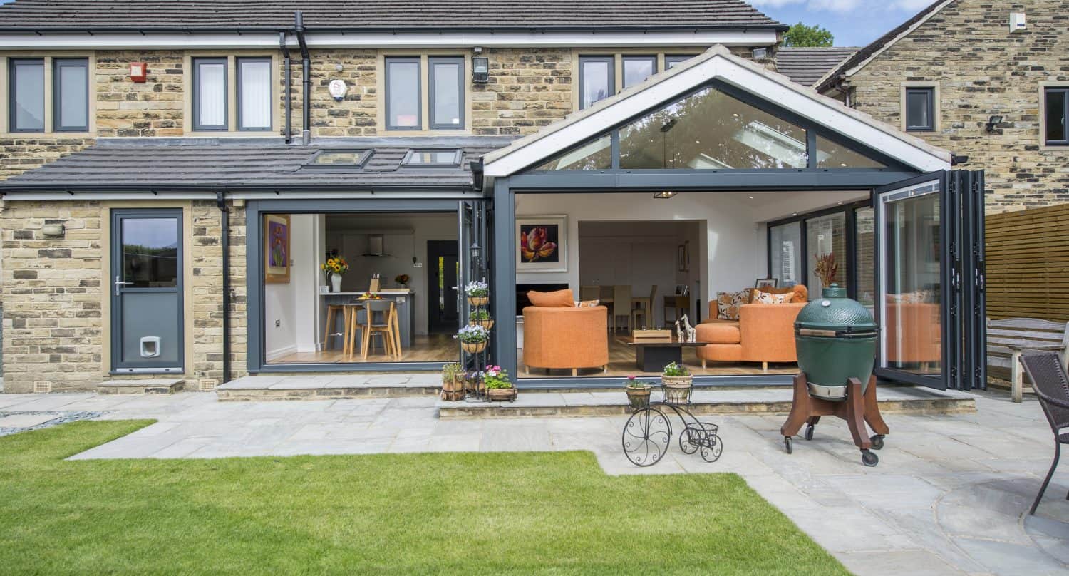 A modern house with a stone exterior features bifold doors that open to a bright living area with orange sofas. A green barbecue grill and potted plants adorn the patio, while the foreground boasts a well-kept lawn.