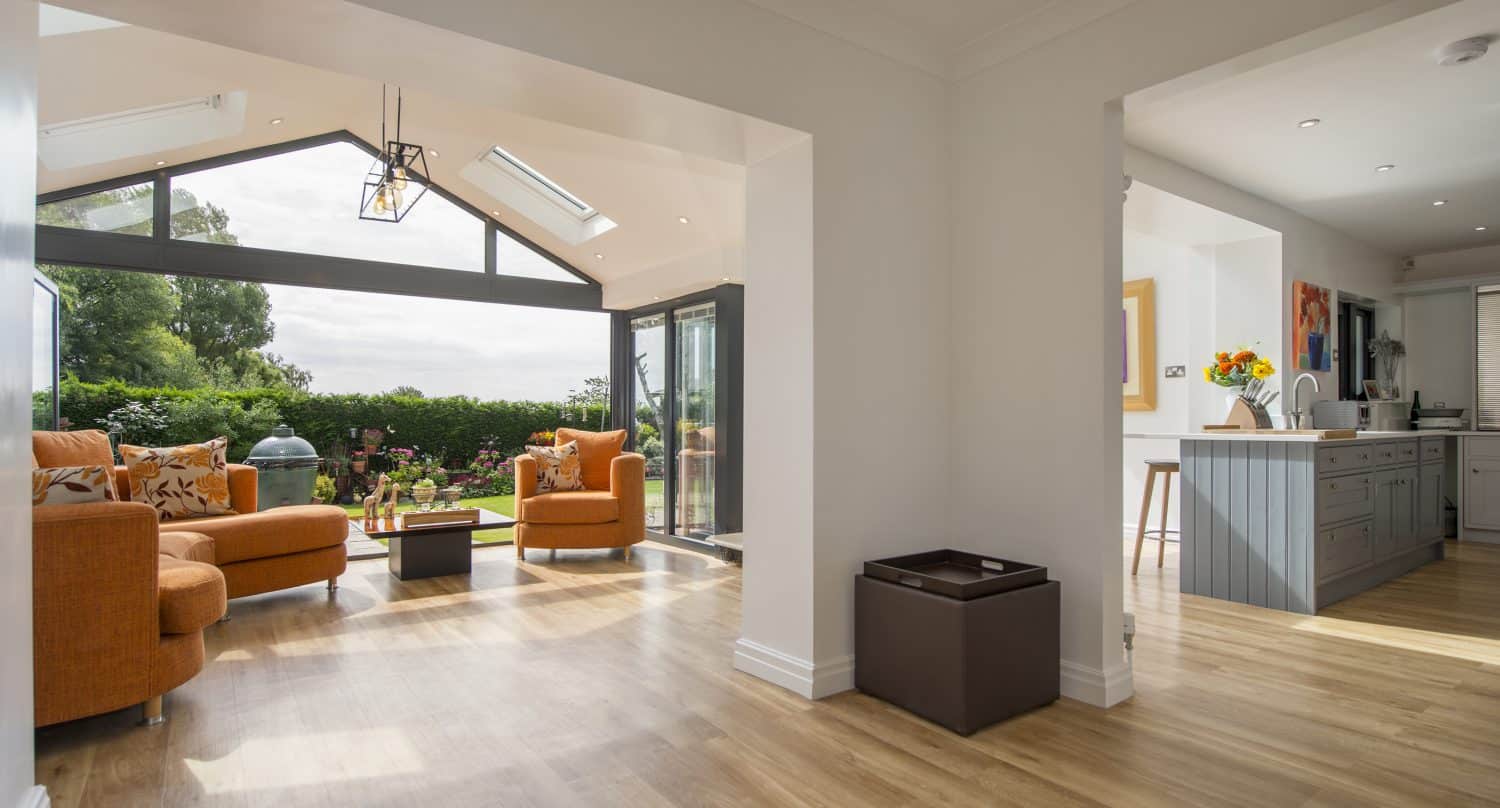 Bright, modern living space with large windows overlooking a garden, orange sofas, a coffee table, light wooden floors, and an open-plan kitchen with a blue island—enhanced by bespoke glass solutions and stylish glass roofing.