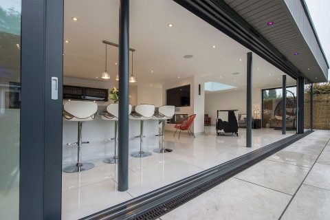 Modern open-plan kitchen and living area with white bar stools, pendant lights, tiled floor, and bespoke glass solutions like sliding doors, viewed from outside on a patio.