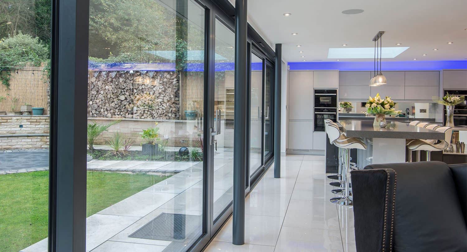 Modern kitchen and dining area with sliding doors and glass walls overlooking a green garden, featuring a sleek island with bar stools, pendant lighting, and stacked firewood outside.