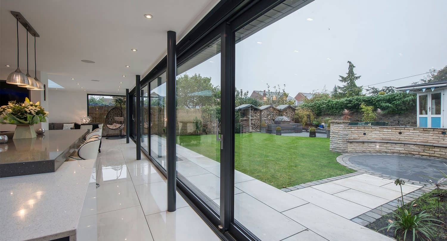 Modern kitchen with large sliding glass doors opening to a landscaped backyard featuring a lawn, stone patio, garden beds, and outdoor seating area. The space is bright and open with contemporary finishes and elegant bifold doors.