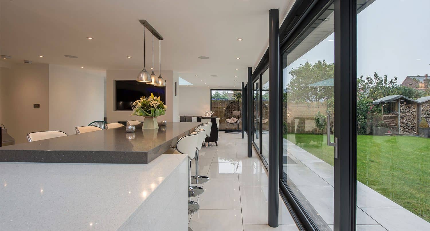 Modern kitchen with white countertops and bar stools, pendant lights above an island, large sliding doors opening to a green backyard, and sleek, minimalistic decor.