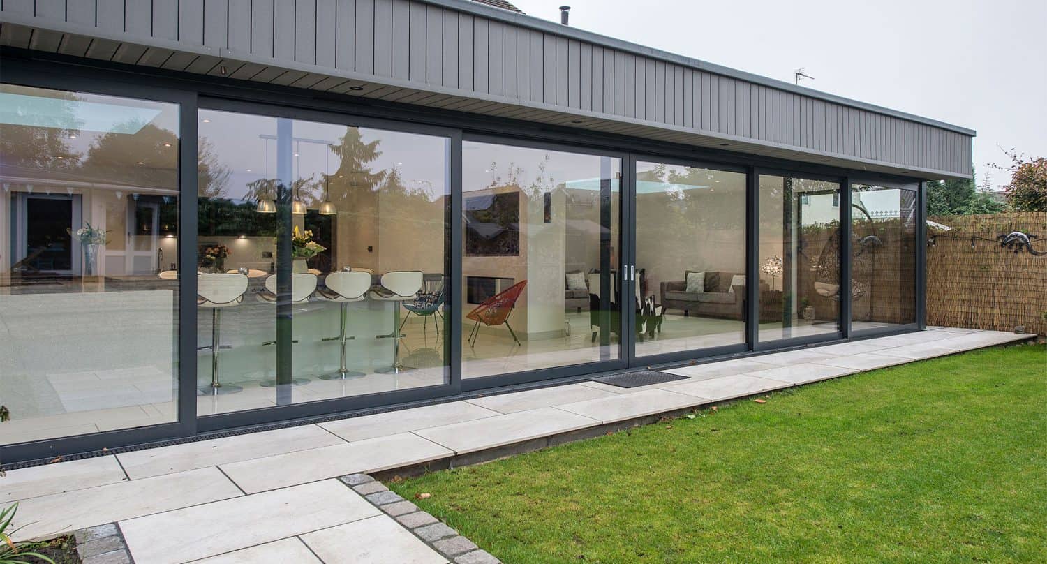 Modern house with large glass sliding doors and bespoke glass solutions, including a sleek glass roofing feature. Inside, a dining table with white chairs and a contemporary living space are visible through the windows.