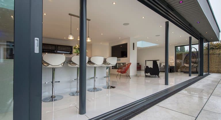 Modern open-plan kitchen and living space with white tile flooring, a row of bar stools at a counter, pendant lights, and large bifold doors opening onto a patio.