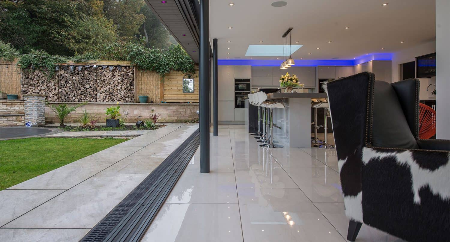 Modern kitchen with glossy tile floors, an island with white barstools, cowhide armchair, and large glass doors open to a patio and green garden. Blue LED ceiling lighting enhances the space, complemented by bespoke glass solutions throughout.