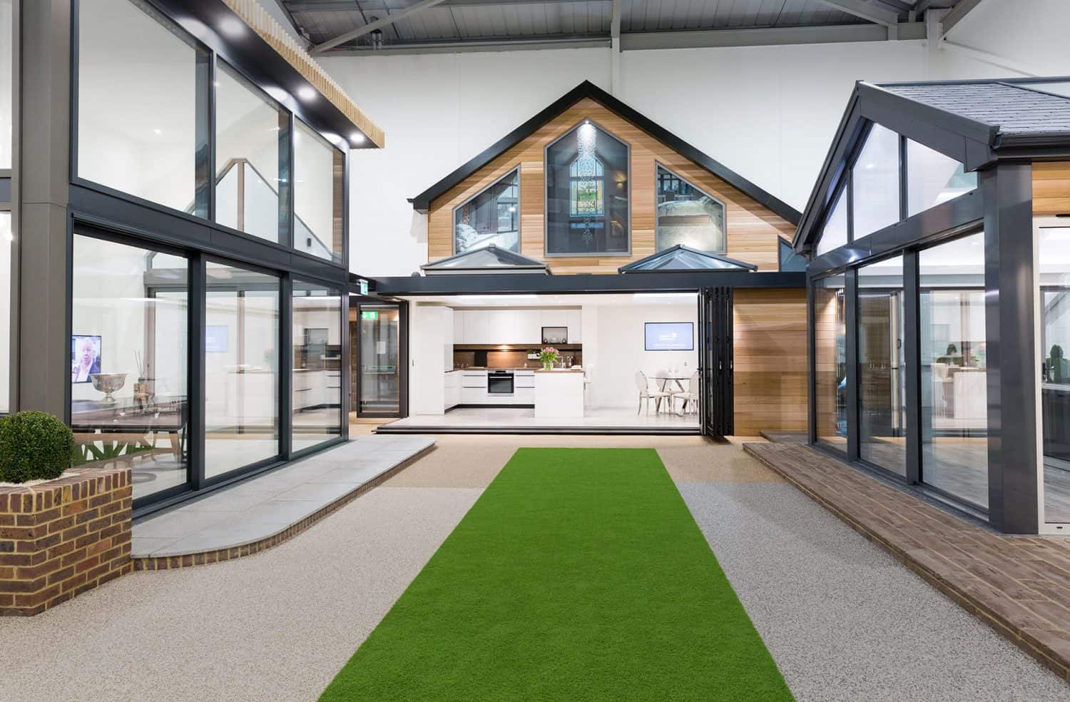 Modern showroom interior featuring a model house with large glass windows, wooden accents, and a green artificial grass walkway leading to a bright kitchen and dining area.