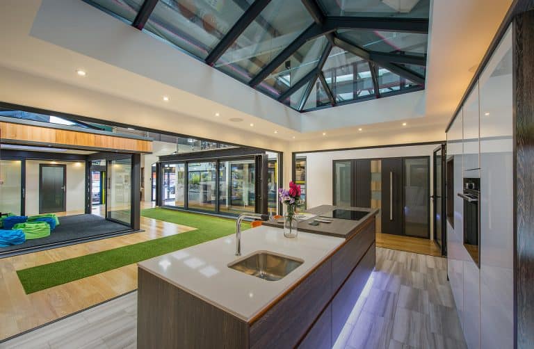 Modern kitchen with a large island, white countertops, and built-in appliances. Bespoke glass solutions shine in the glass ceiling panels, while open spaces and a mix of wooden and green artificial turf flooring add unique flair.