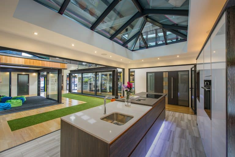 Modern kitchen with a large island, white countertops, and built-in appliances. Bespoke glass solutions shine in the glass ceiling panels, while open spaces and a mix of wooden and green artificial turf flooring add unique flair.
