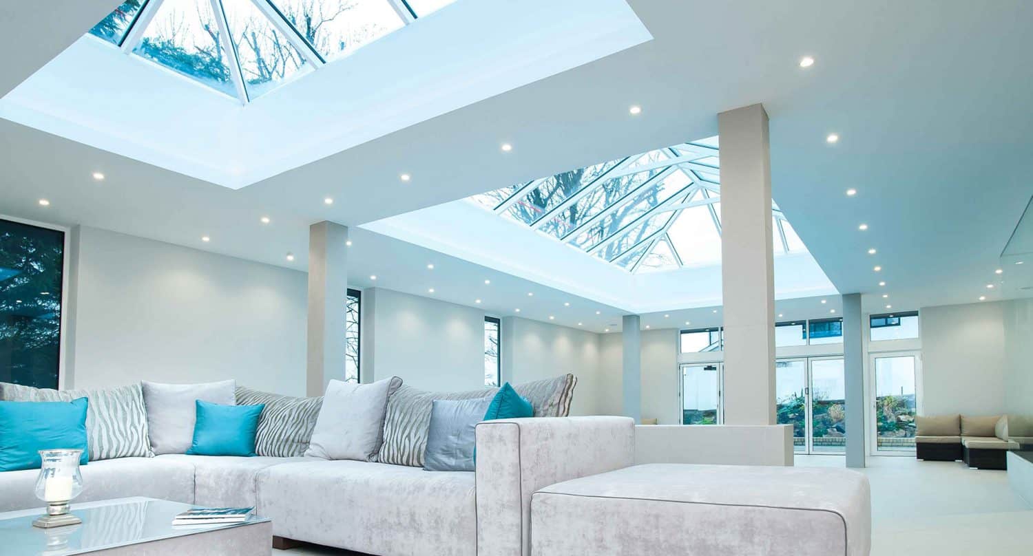 A modern, spacious living room with white walls, large skylights, recessed lighting, and contemporary white and grey furniture accented with blue and striped pillows. Trees are visible through the skylights and windows.