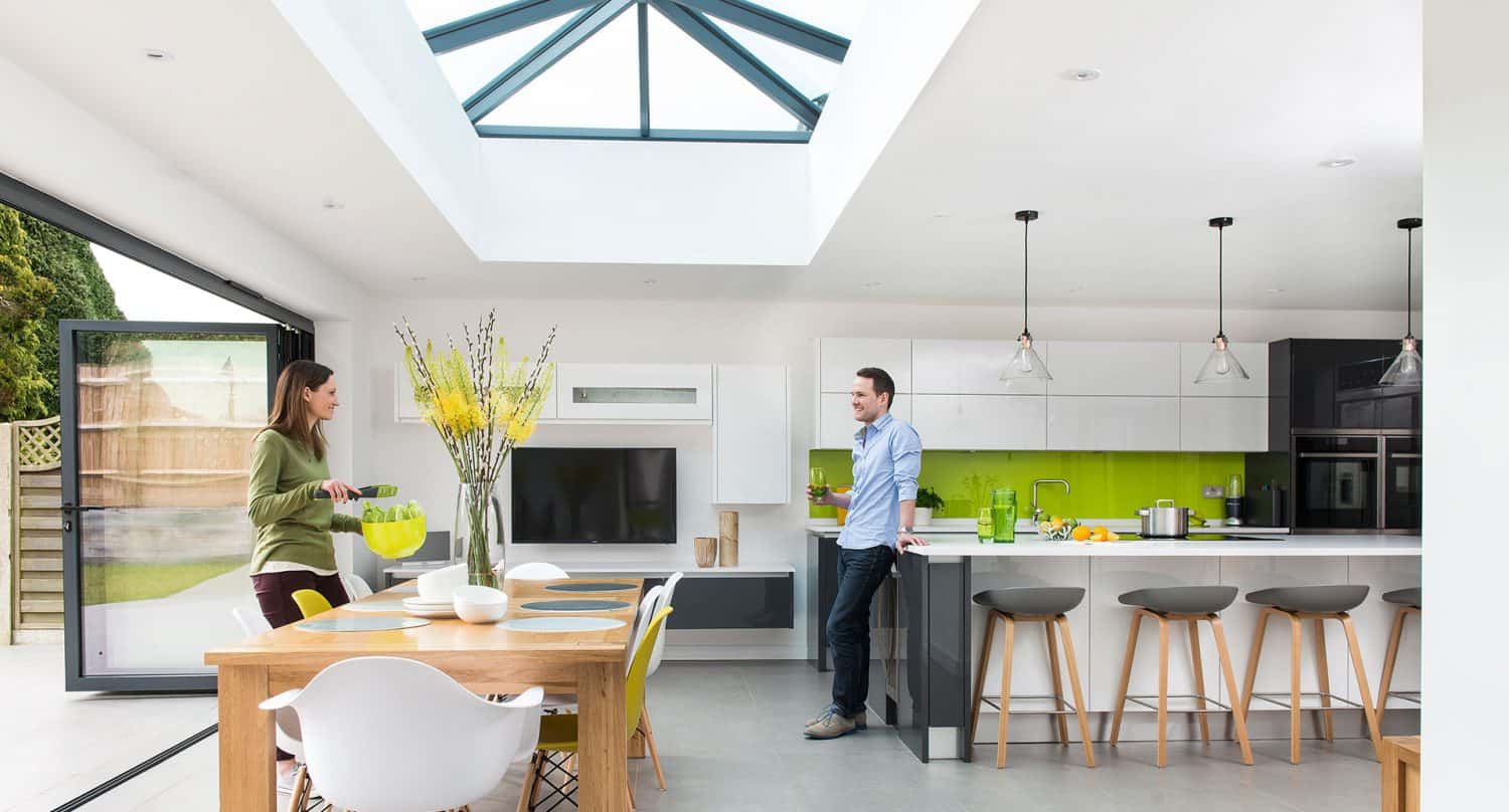 A modern, bright kitchen with a skylight, white cabinets, green backsplash, and an island with stools. A woman pours a drink at a wooden dining table while a man leans against the kitchen counter, both smiling.