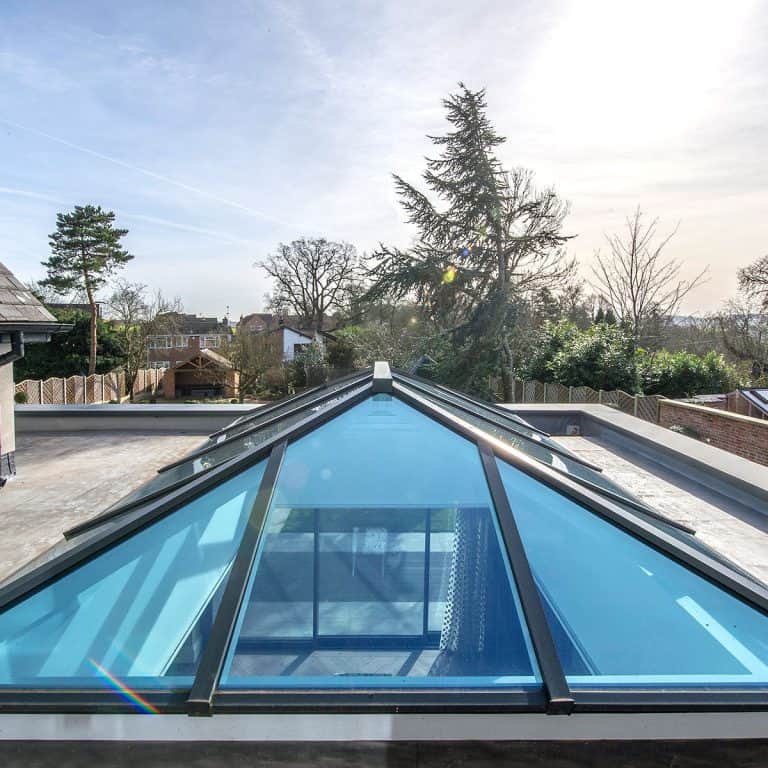 A modern glass roof lantern with a metal frame on a flat rooftop, reflecting the sky and surrounding trees on a bright, sunny day. Houses and trees are visible in the background.