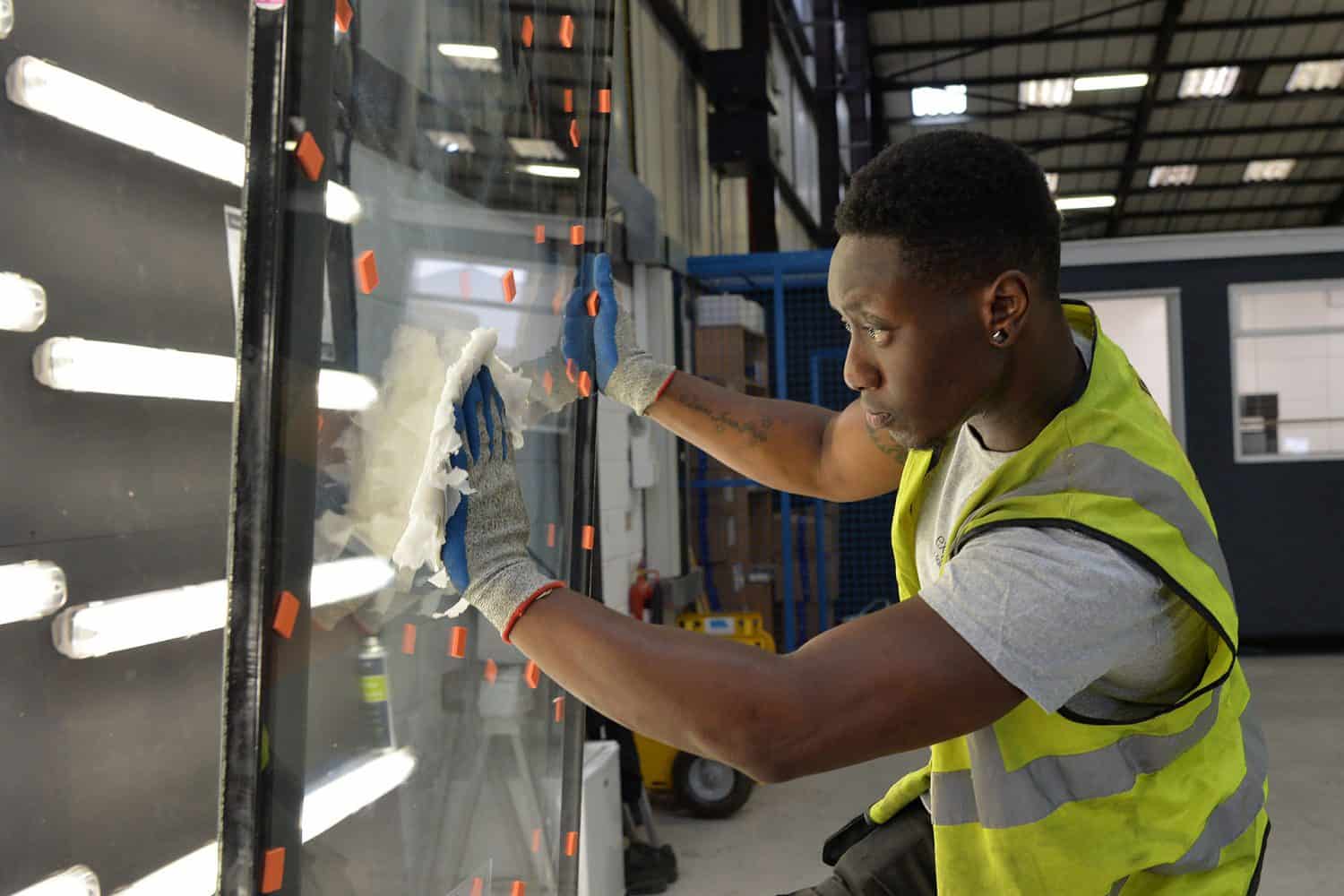 A person wearing a high-visibility vest and gloves is cleaning or inspecting a large glass panel in an industrial setting with bright overhead lighting.
