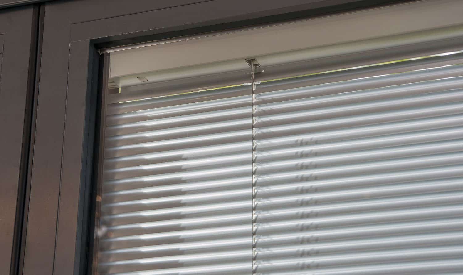 Close-up view of a window with horizontal metallic blinds, partially open, set in a modern dark-colored frame. Light is softly filtering through the slats from outside.