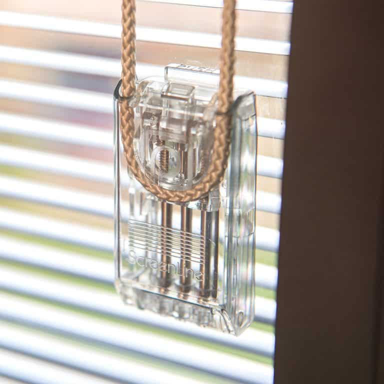 A clear plastic window blind cord safety device is attached to a beaded cord in front of horizontal window blinds with sunlight streaming through.