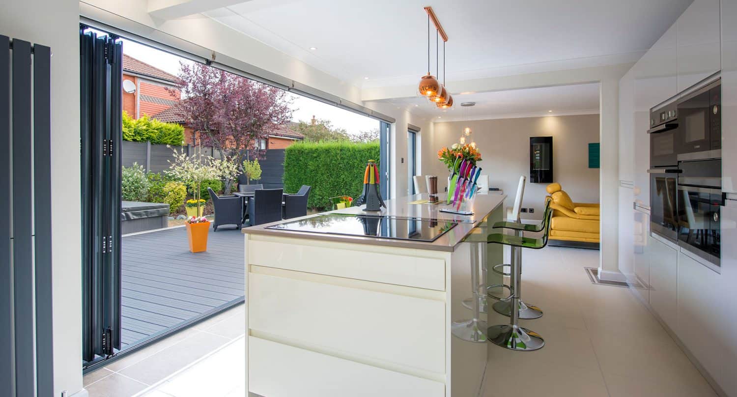 Modern kitchen with white island, built-in appliances, and pendant lights, opening onto a patio with outdoor seating and greenery through large folding glass doors.