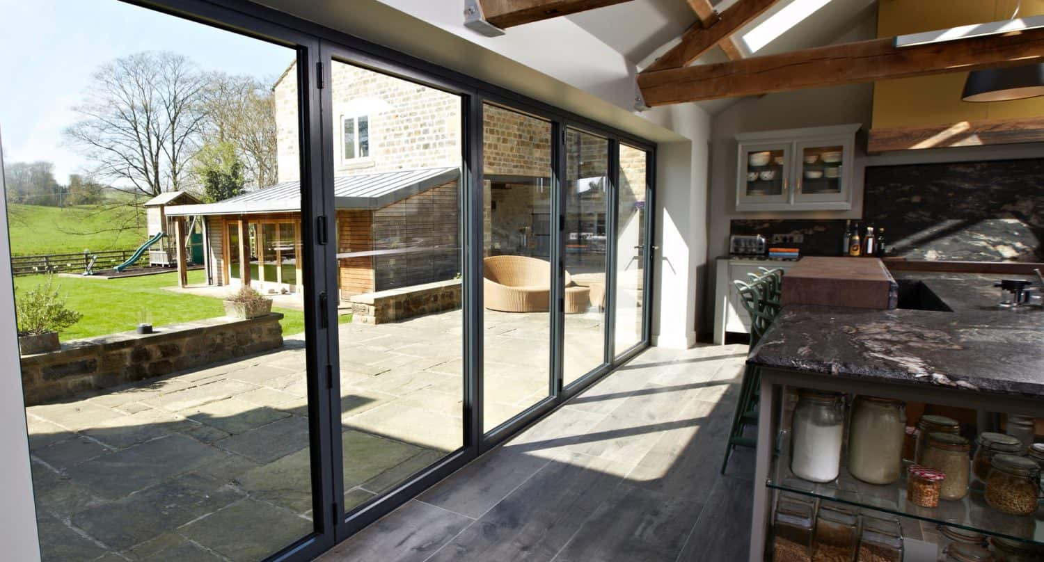 Modern kitchen with large glass sliding doors opening onto a sunny stone patio and green backyard; wooden beams on the ceiling and glass jars on the kitchen counter are visible.